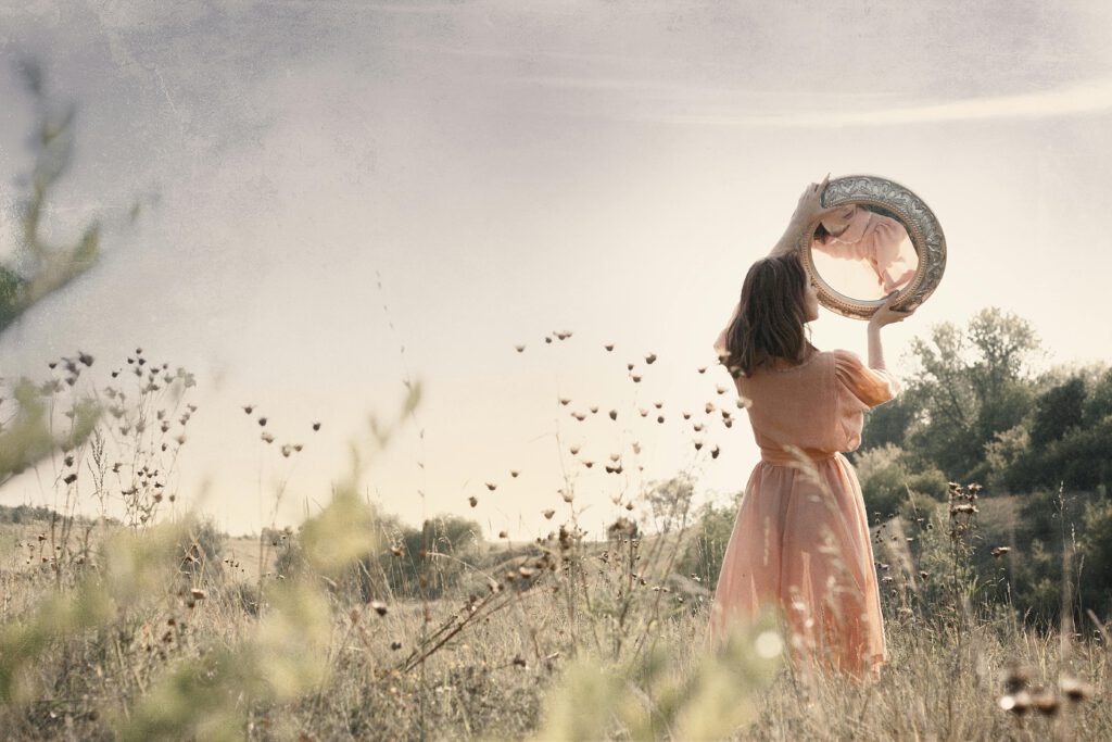 Young woman in a field holds a mirror, reflecting a serene outdoor scene in natural light.